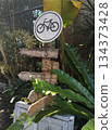 A wooden bicycle sign in a garden, with arrows indicating directions. Green plants surround the sign, creating a natural scene 134373428