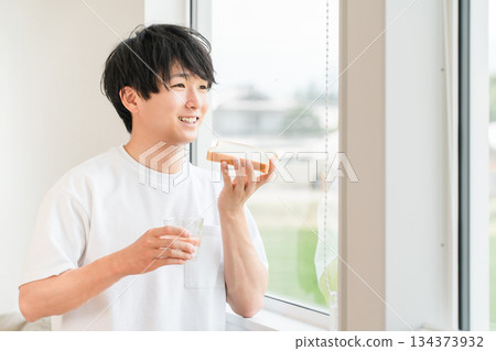 一位年輕的亞洲男子在他家窗邊吃麵包 134373932