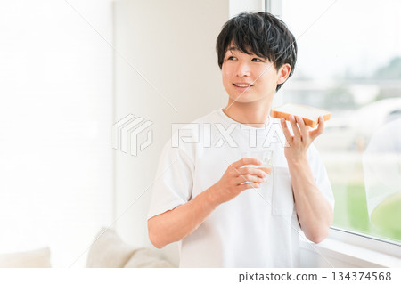 一位年輕的亞洲男子在他家窗邊吃麵包 134374568