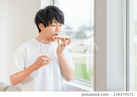 A young Asian man eating bread by the window in his home 134374571
