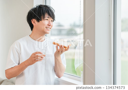 一位年輕的亞洲男子在他家窗邊吃麵包 一位年輕的亞洲男子在他家窗邊吃麵包 134374573