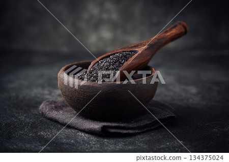Black lava salt in rustic wooden bowl with matching spoon on dark textured background creates dramatic and elegant culinary presentation for gourmet seasoning 134375024