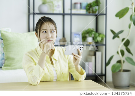 Portrait of a thinking Japanese woman watching videos on her smartphone in the living room Portrait of a thinking Japanese woman watching videos on her smartphone in the living room 134375724