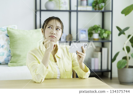 Portrait of a thinking Japanese woman watching videos on her smartphone in the living room 134375744