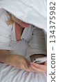 Smiling woman lying on a bed with a blanket over her head while using the mobile phone 134375982