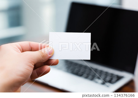 Business card in hand on laptop background. Hand holding blank business card in front of laptop. Blank white business card mockup in office environment. Business card in hand on laptop background. Hand holding blank business card in front of laptop. Blank white business card mockup in office environment. 134376166