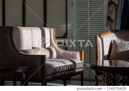 Sunlight shines on the elegant wooden sofa upholstered with ivory velvet fabric and a round coffee table set in a living room. Sunlight shines on the elegant wooden sofa upholstered with ivory velvet fabric and a round coffee table set in a living room. 134378428