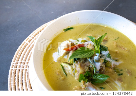 Close up to Green Curry with Chicken garnished on top with pepper and basil was served with white bowl put on wicker cream color placemat. 134378442
