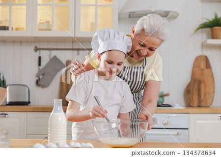 Happy family in kitchen. Grandmother and granddaughter child cook in kitchen together. Grandma teaching kid girl knead dough bake cookies. Household teamwork helping family generations concept 134378634