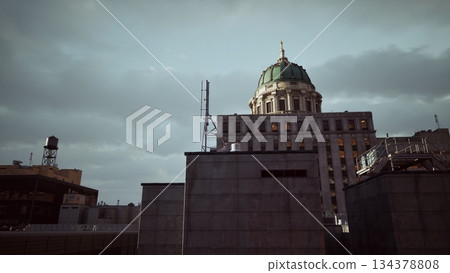 As twilight falls over the city, the elegant dome of a historic building stands out against the cloudy sky, surrounded by modern rooftops and urban structures, evoking a sense of timelessness. 134378808