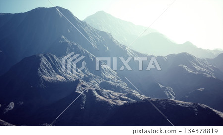 Towering mountains stretch across the horizon bathed in golden light. The rugged terrain displays unique textures and shades, creating a breathtaking and tranquil atmosphere. Towering mountains stretch across the horizon bathed in golden light. The rugged terrain displays unique textures and shades, creating a breathtaking and tranquil atmosphere. 134378819