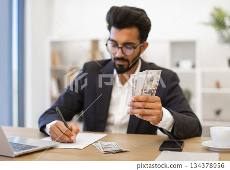 An Indian businessman is working at his desk, counting money and making notes An Indian businessman is working at his desk, counting money and making notes 134378956