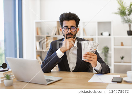 An Indian businessman in a suit holds a wad of cash while working in his office 134378972