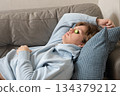 Young man relax with cucumber slices on eyes, doing simple skincare and relaxing indoors in cozy home interior. Leisure Activities At Home 134379212
