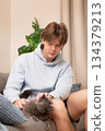Young man sitting on sofa and gently petting grey one eyed british cat indoors, showing calm interaction, care and bonding in cozy home setting. Leisure Activities At Home 134379213