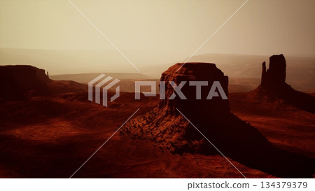 At dusk, warm hues illuminate the vast desert landscape of Monument Valley, highlighting towering red rock formations. Shadows stretch across the sandy terrain, creating a serene atmosphere. 134379379