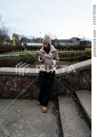 Chilly afternoon in the park as a young woman checks her phone while dressed warmly in a cozy jacket and hat, surrounded by tranquil nature and muted skies 134379566