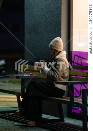 Evening solitude on a city bench with a warm drink, where moments of reflection come alive against the glow of urban lights 134379568