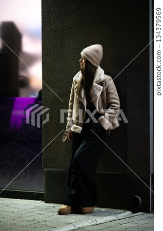 Fashionable young woman in cozy winter attire leans against a stylish city wall during twilight hours, capturing the essence of urban chic 134379569