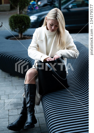 Dressed in elegance, a woman with long blonde hair sits on a curved bench in a modern urban setting while preparing to retrieve something from her bag 134379577