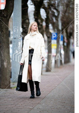 Stylish young woman strolls through a city street in winter attire, showcasing a blend of modern fashion and comfort during the early afternoon hours Stylish young woman strolls through a city street in winter attire, showcasing a blend of modern fashion and comfort during the early afternoon hours 134379580