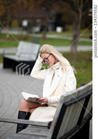 Reading in tranquility, a young woman enjoys a quiet afternoon in a park, lost in the pages of a book while seated on a weathered bench surrounded by autumn foliage Reading in tranquility, a young woman enjoys a quiet afternoon in a park, lost in the pages of a book while seated on a weathered bench surrounded by autumn foliage 134379582