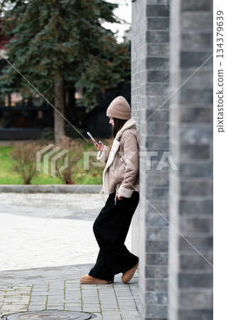 Fashionable woman using smartphone while leaning against a stone wall in a modern urban setting with greenery in the background during a chilly afternoon Fashionable woman using smartphone while leaning against a stone wall in a modern urban setting with greenery in the background during a chilly afternoon 134379639