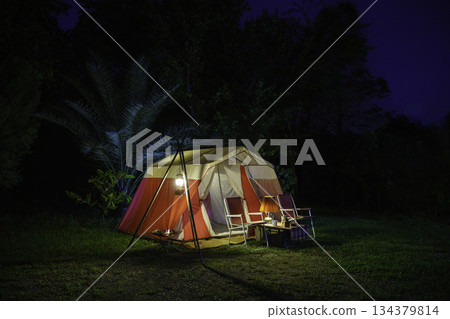 Camping tents at night, vintage cabin tents, antique oil lamps, retro chairs, and a group of camping tents with outdoor coffee-making facilities on wooden tables in a forest camping area. 134379814