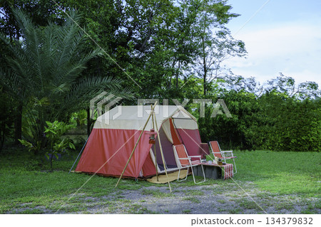 camping setup in the evening, vintage cabin tents, antique oil lamps, retro chairs, and a group of camping tents with outdoor coffee-making facilities on tables in a forest camping area. 134379832