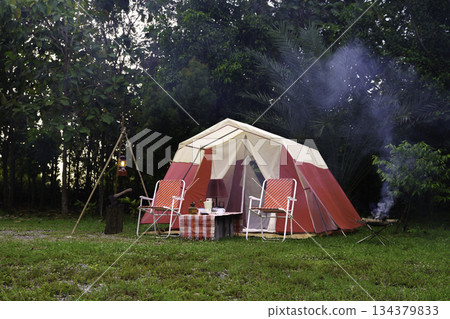 camping setup in the evening, vintage cabin tents, antique oil lamps, retro chairs, and a group of camping tents with outdoor coffee-making facilities on tables in a forest camping area. 134379833