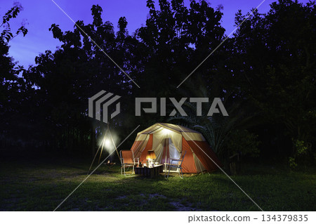 Camping tents at night, vintage cabin tents, antique oil lamps, retro chairs, and a group of camping tents with outdoor coffee-making facilities on wooden tables in a forest camping area. Camping tents at night, vintage cabin tents, antique oil lamps, retro chairs, and a group of camping tents with outdoor coffee-making facilities on wooden tables in a forest camping area. 134379835