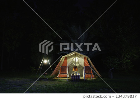 Camping tents at night, vintage cabin tents, antique oil lamps, retro chairs, and a group of camping tents with outdoor coffee-making facilities on wooden tables in a forest camping area. Camping tents at night, vintage cabin tents, antique oil lamps, retro chairs, and a group of camping tents with outdoor coffee-making facilities on wooden tables in a forest camping area. 134379837