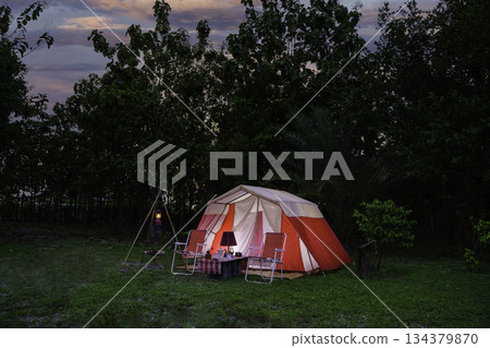 Camping tents at night, vintage cabin tents, antique oil lamps, retro chairs, and a group of camping tents with outdoor coffee-making facilities on wooden tables in a forest camping area. 134379870