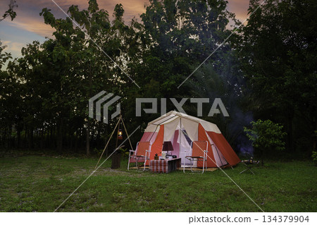 Camping tents at night, vintage cabin tents, antique oil lamps, retro chairs, and a group of camping tents with outdoor coffee-making facilities on wooden tables in a forest camping area. 134379904