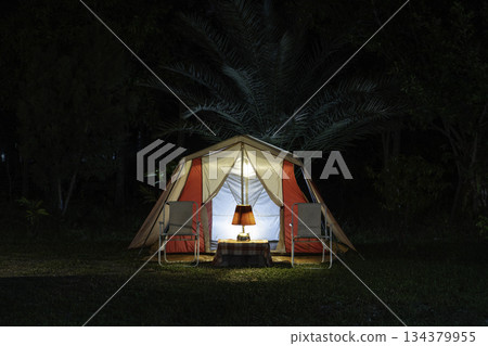 Camping tents at night, vintage cabin tents, antique oil lamps, retro chairs, and a group of camping tents with outdoor coffee-making facilities on wooden tables in a forest camping area. Camping tents at night, vintage cabin tents, antique oil lamps, retro chairs, and a group of camping tents with outdoor coffee-making facilities on wooden tables in a forest camping area. 134379955