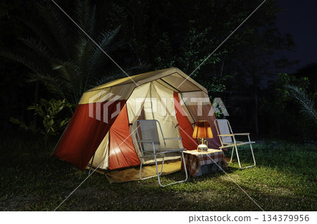 Camping tents at night, vintage cabin tents, antique oil lamps, retro chairs, and a group of camping tents with outdoor coffee-making facilities on wooden tables in a forest camping area. Camping tents at night, vintage cabin tents, antique oil lamps, retro chairs, and a group of camping tents with outdoor coffee-making facilities on wooden tables in a forest camping area. 134379956