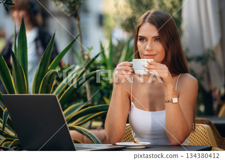 Fresh hot drink in cup. Beautiful young woman is outdoors of the restaurant by the table 134380412