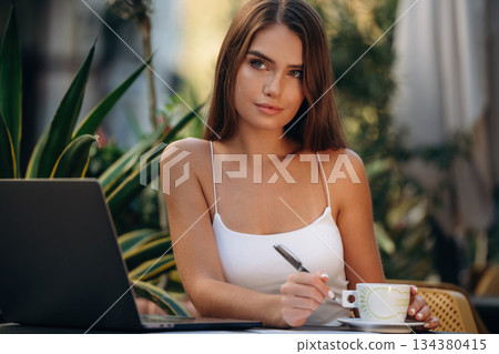 Business deals, with laptop and pen. Beautiful young woman is outdoors of the restaurant by the table Business deals, with laptop and pen. Beautiful young woman is outdoors of the restaurant by the table 134380415