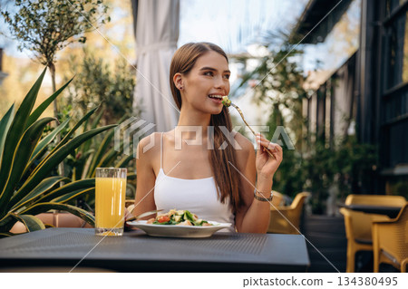 Eating food that is on the plate. Beautiful young woman is outdoors of the restaurant by the table Eating food that is on the plate. Beautiful young woman is outdoors of the restaurant by the table 134380495
