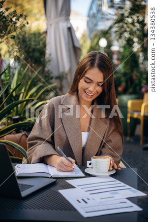 Remote work, with drink and documents. Beautiful young woman is outdoors of the restaurant by the table Remote work, with drink and documents. Beautiful young woman is outdoors of the restaurant by the table 134380538