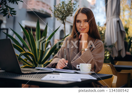 Remote work, with drink and documents. Beautiful young woman is outdoors of the restaurant by the table Remote work, with drink and documents. Beautiful young woman is outdoors of the restaurant by the table 134380542