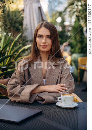 Fresh hot drink in cup. Beautiful young woman is outdoors of the restaurant by the table 134380547