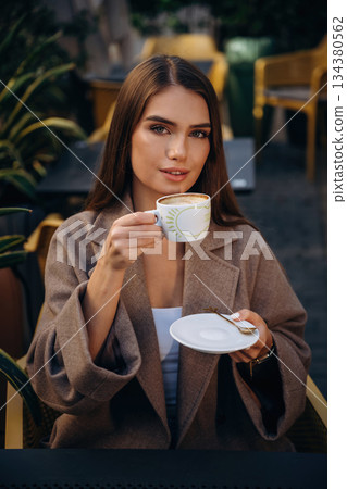 Looking at the camera, sitting. Beautiful young woman is outdoors of the restaurant by the table Looking at the camera, sitting. Beautiful young woman is outdoors of the restaurant by the table 134380562