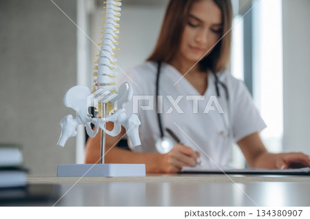 Writing in the document, spine model is on the table. Professional female doctor in the clinic indoors 134380907