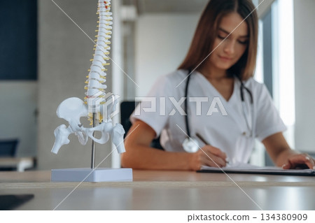 Writing in the document, spine model is on the table. Professional female doctor in the clinic indoors Writing in the document, spine model is on the table. Professional female doctor in the clinic indoors 134380909