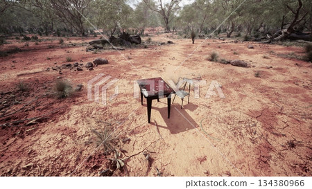 A solitary table and chair stand on cracked earth surrounded by sparse vegetation. The scene captures the desolation of a dry landscape, evoking a sense of isolation and stillness. 134380966