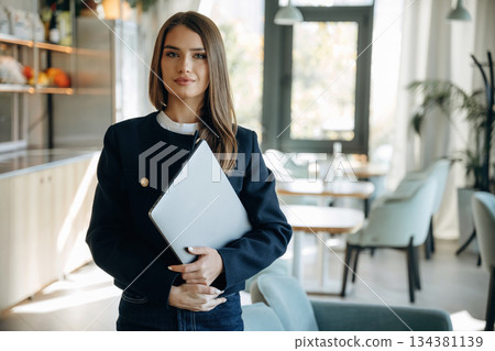 Businesswoman is standing indoors and holding laptop Businesswoman is standing indoors and holding laptop 134381139