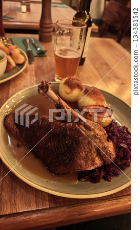 Roasted goose leg with crispy skin served with red cabbage, potato dumplings and baked apple on a restaurant plate. Concept of traditional comfort food, hearty cuisine and classic European dining Roasted goose leg with crispy skin served with red cabbage, potato dumplings and baked apple on a restaurant plate. Concept of traditional comfort food, hearty cuisine and classic European dining 134381542