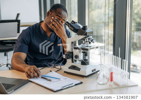 Tired, sitting and taking a break. Scientist is working indoors, microscope is on the table Tired, sitting and taking a break. Scientist is working indoors, microscope is on the table 134381907