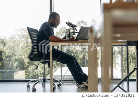 Side view, sitting. Scientist is working indoors, microscope is on the table Side view, sitting. Scientist is working indoors, microscope is on the table 134381921